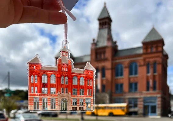 A photograph of a hand holding an illustrated acrylic ornament in front of a large brick building.